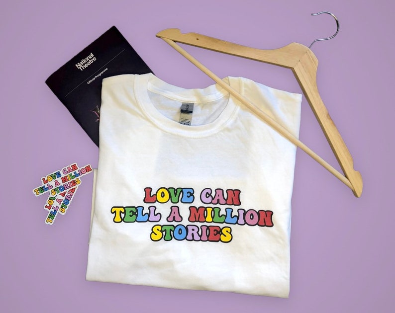 May include: White t-shirt with the text "LOVE CAN TELL A MILLION STORIES" in rainbow letters. The shirt is folded and on a purple surface with a wooden hanger and a National Theatre program.
