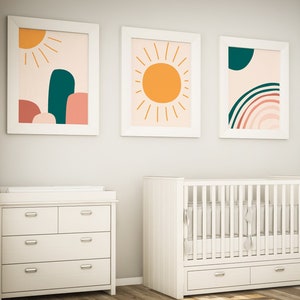 May include: A nursery room with a white crib, dresser, and rocking chair. Three framed abstract art prints hang on the wall, featuring sun and rainbow designs in orange, teal, and pink. The room has a window with white curtains.