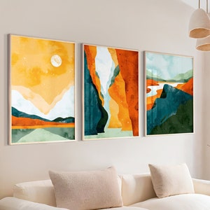 May include: Three abstract landscape paintings in a living room with a white couch, a brown armchair, and a white coffee table. The paintings are in shades of yellow, orange, green, and blue.