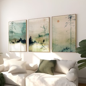 May include: Three framed watercolour paintings depicting a misty forest scene with trees, a bird, and a red sun. The paintings are hung on a white wall above a white sofa with green throw pillows and a green throw blanket.