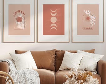 May include: Three framed art prints with a celestial theme hang above a brown sofa. Each print features a different design in shades of peach and white, including suns, moons, and botanical elements. The room is decorated in neutral tones with a modern aesthetic.