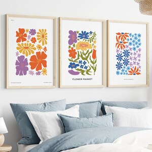 K&ouml;nnte beinhalten: Drei gerahmte Drucke mit bunten Blumenmustern. Die Drucke sind in einer Reihe an einer wei&szlig;en Wand angeordnet. Die Drucke zeigen verschiedene Farben und Muster von Blumen, darunter Orange, Gelb, Blau, Lila und Gr&uuml;n.