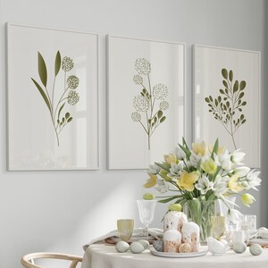 May include: A white tablecloth with a floral centrepiece of yellow and white flowers in a clear glass vase. The table is set with two wooden chairs, two white plates, and two glasses. There are three framed prints on the wall behind the table, each featuring a green botanical illustration.