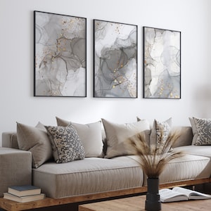 May include: A light gray sectional sofa with a wooden frame and a light gray throw pillow with a geometric pattern. The sofa is in front of a white wall with three abstract paintings in black and gray with gold accents. A wooden coffee table sits in front of the sofa.