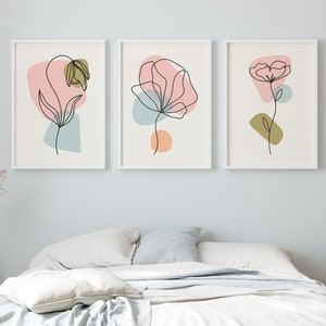 May include: Three minimalist line art prints featuring abstract flowers with pink, blue, and green accents. The prints are framed in white and hung on a light blue wall above a white bed with a white throw blanket.