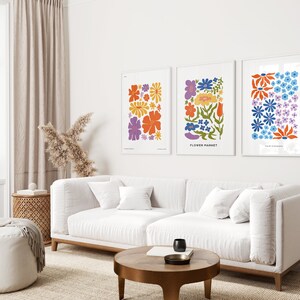 K&ouml;nnte beinhalten: Drei gerahmte Drucke mit bunten Blumenillustrationen. Die Drucke h&auml;ngen an einer wei&szlig;en Wand &uuml;ber einem wei&szlig;en Sofa. Der mittlere Druck hat den Text "FLOWER MARKET" unten.