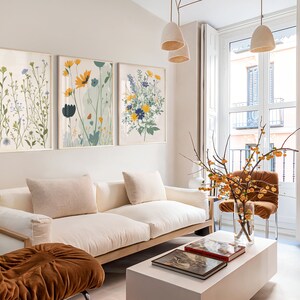May include: Three framed floral art prints with gold frames hang above a white sofa. The prints feature botanical illustrations in shades of green, yellow, blue, and white. The room has a modern, minimalist aesthetic with neutral tones.