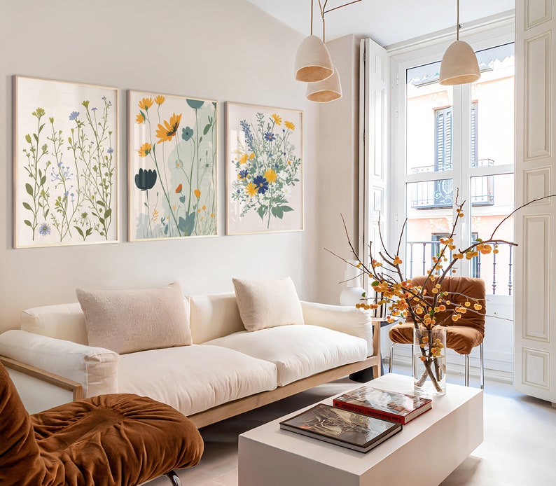 May include: Three framed floral art prints with gold frames hang above a white sofa. The prints feature botanical illustrations in shades of green, yellow, blue, and white. The room has a modern, minimalist aesthetic with neutral tones.