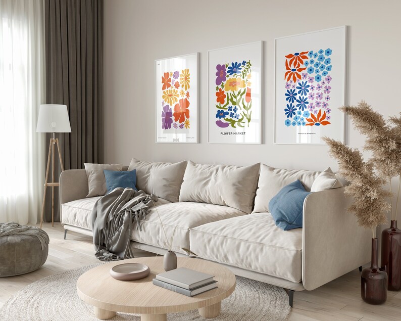 K&ouml;nnte beinhalten: Drei gerahmte Blumenprints mit bunten abstrakten Blumen in Orange-, Gelb-, Blau-, Lila- und Gr&uuml;nt&ouml;nen. Die Drucke h&auml;ngen an einer hellbeigen Wand &uuml;ber einem wei&szlig;en Sofa.