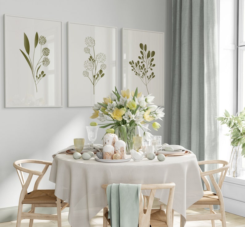 May include: A white tablecloth with a floral centrepiece of yellow and white flowers in a clear glass vase. The table is set with two wooden chairs, two white plates, and two glasses. There are three framed prints on the wall behind the table, each featuring a green botanical illustration.