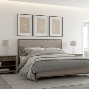 May include: A gray upholstered bed with a wooden frame and a gray duvet cover. The bed is in a bedroom with white walls and a rug on the floor. There are two bedside tables with lamps on them and three framed prints on the wall above the headboard.