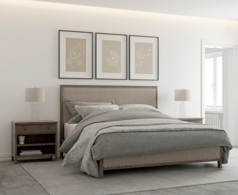 May include: A gray upholstered bed with a wooden frame and a gray duvet cover. The bed is in a bedroom with white walls and a rug on the floor. There are two bedside tables with lamps on them and three framed prints on the wall above the headboard.