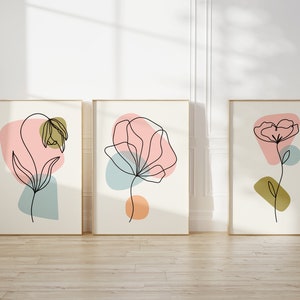 May include: Three framed prints with abstract line drawings of flowers in various colors, including pink, blue, and green. The prints are set against a white wall.