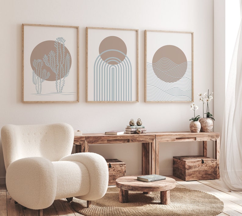 May include: Three minimalist art prints with a light brown background and blue geometric designs. The prints are framed in natural wood and hung on a white wall above a wooden table.