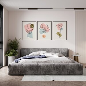 May include: Three framed prints with abstract line drawings of flowers in pink, orange, and green hues on a white background. The prints are hanging on a white wall above a gray upholstered bed.