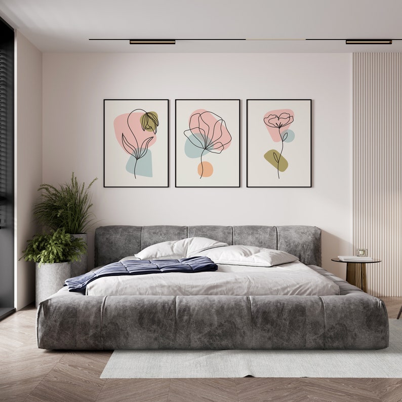 May include: Three framed prints with abstract line drawings of flowers in pink, orange, and green hues on a white background. The prints are hanging on a white wall above a gray upholstered bed.