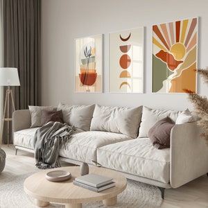 May include: A living room interior with a white sofa, a round wooden coffee table, a grey throw blanket, and three abstract wall art prints with geometric shapes and colours. The prints feature a sun, moon, and a stack of bowls.
