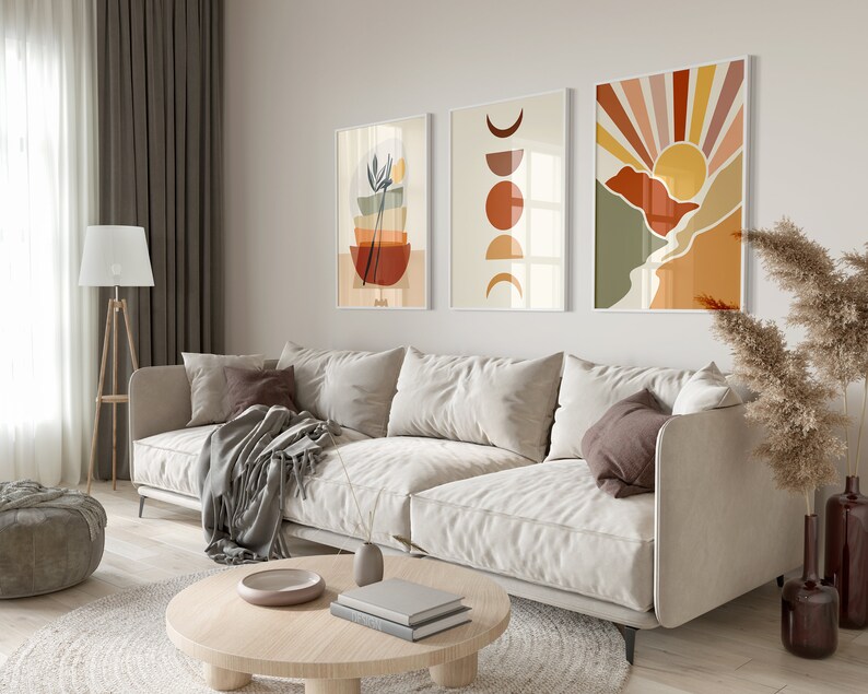 May include: A living room interior with a white sofa, a round wooden coffee table, a grey throw blanket, and three abstract wall art prints with geometric shapes and colours. The prints feature a sun, moon, and a stack of bowls.