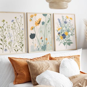 May include: Three framed floral art prints. The prints feature botanical illustrations of wildflowers in shades of green, yellow, blue, and orange. The frames are a light wood color. The art is displayed above a bed with orange and tan pillows.