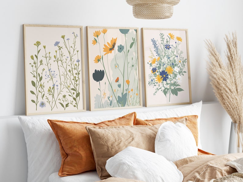 May include: Three framed floral art prints. The prints feature botanical illustrations of wildflowers in shades of green, yellow, blue, and orange. The frames are a light wood color. The art is displayed above a bed with orange and tan pillows.
