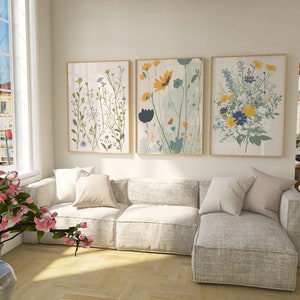 May include: Three framed floral art prints with botanical illustrations in a living room setting. The prints feature colorful flowers in shades of yellow, blue, and green. A light gray sectional sofa with pillows is in front of the art.
