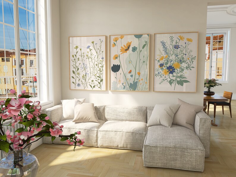 May include: Three framed floral art prints with botanical illustrations in a living room setting. The prints feature colorful flowers in shades of yellow, blue, and green. A light gray sectional sofa with pillows is in front of the art.