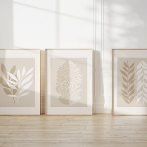 May include: Three framed prints with minimalist beige and white botanical illustrations. The prints are in gold frames and are hanging on a white wall.
