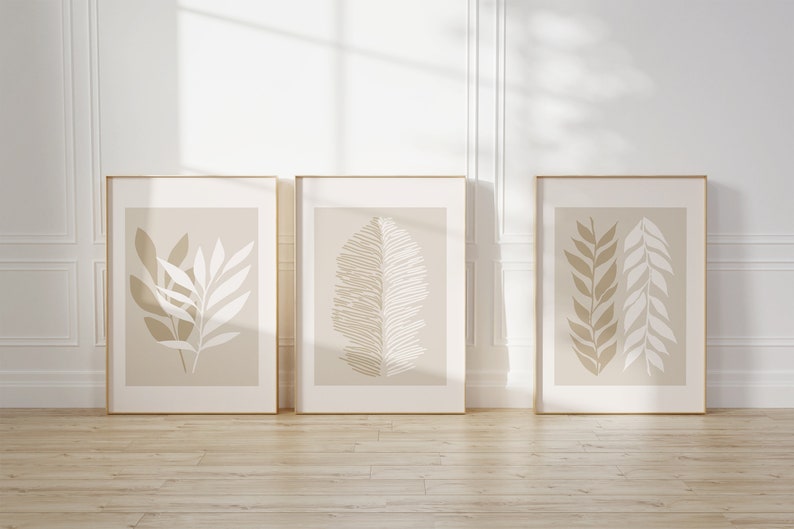May include: Three framed prints with minimalist beige and white botanical illustrations. The prints are in gold frames and are hanging on a white wall.
