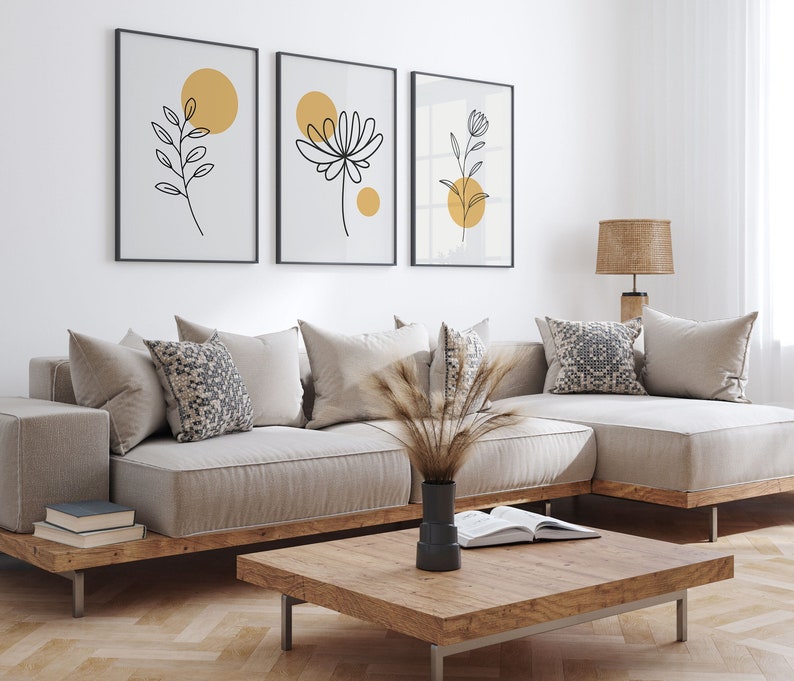 May include: A beige sectional sofa with a wooden frame and a coffee table in front of it. The sofa has three pillows, one with a geometric pattern. Three framed prints with black line drawings of flowers and circles in yellow are hanging on the wall behind the sofa.