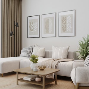 May include: A white sectional sofa with three throw pillows, a throw blanket, and a coffee table with a book and decorative items. The sofa is in a living room with a large window, beige curtains, and three framed prints with leaf designs on the wall.