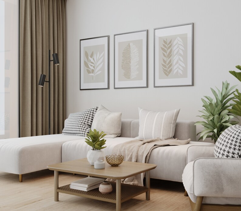 May include: A white sectional sofa with three throw pillows, a throw blanket, and a coffee table with a book and decorative items. The sofa is in a living room with a large window, beige curtains, and three framed prints with leaf designs on the wall.