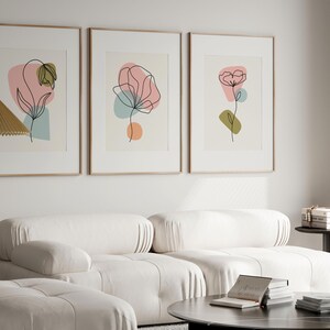May include: Three framed prints with abstract line drawings of flowers in pink, green, and blue colors. The prints are hung on a white wall above a white sofa.