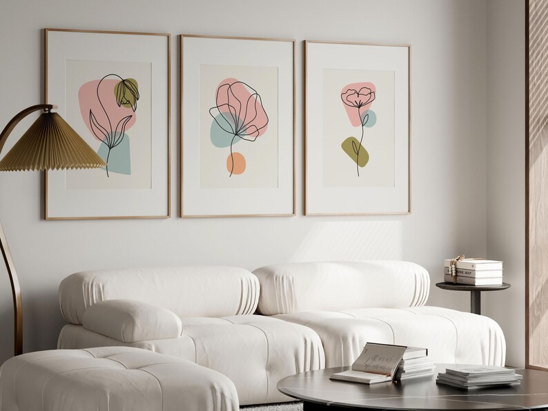 May include: Three framed prints with abstract line drawings of flowers in pink, green, and blue colors. The prints are hung on a white wall above a white sofa.