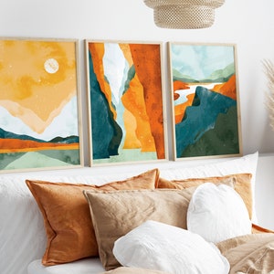May include: Three framed abstract landscape paintings in a minimalist style. The paintings feature a mix of orange, yellow, green, and blue colors. The paintings are hung on a white wall above a bed with pillows.