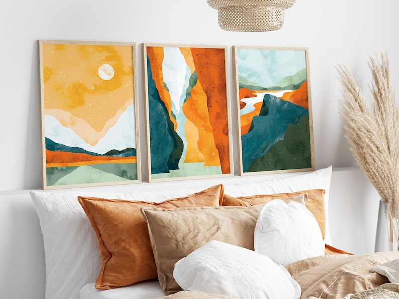 May include: Three framed abstract landscape paintings in a minimalist style. The paintings feature a mix of orange, yellow, green, and blue colors. The paintings are hung on a white wall above a bed with pillows.