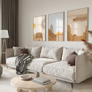 May include: A living room interior with a white sofa, a coffee table, a lamp, and three abstract art prints on the wall. The prints are in shades of brown, beige, and yellow.