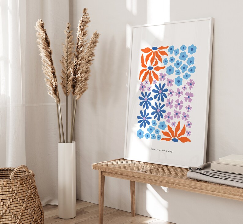 K&ouml;nnte beinhalten: Ein wei&szlig;er gerahmter Druck mit einem farbenfrohen Blumendesign. Der Druck zeigt blaue, orange und lila Blumen auf wei&szlig;em Hintergrund. Der Text "The Art of Simplicity" befindet sich am unteren Rand des Drucks.