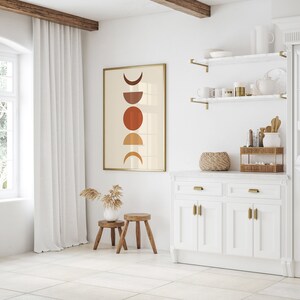 May include: A white kitchen cabinet with two drawers and four doors. The cabinet has gold hardware and a white marble countertop. A framed print with a geometric design of circles and crescent moons in shades of brown and orange hangs on the wall above the cabinet.