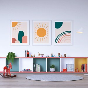 May include: Three framed art prints with abstract designs. One features a sun, another a sun with a mountain-like shape, and the third a rainbow. The prints are displayed above a colorful shelf with toys and books. A red rocking horse is on the floor.