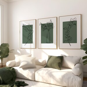 May include: Three framed line drawings of floral arrangements on a white wall. The drawings are in black and white and feature a green background. The drawings are hung above a white sofa with green throw cushions.