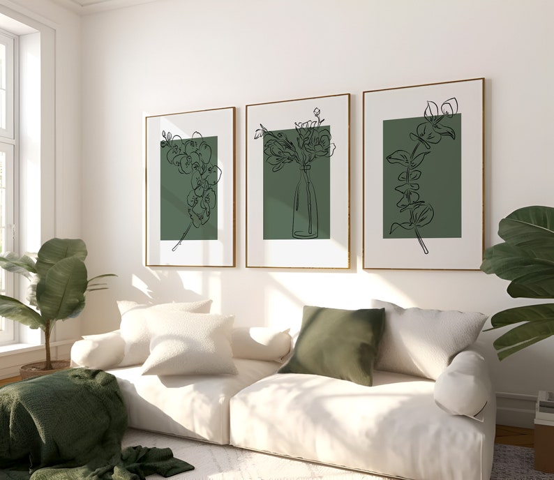 May include: Three framed line drawings of floral arrangements on a white wall. The drawings are in black and white and feature a green background. The drawings are hung above a white sofa with green throw cushions.