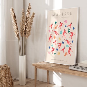 Puede incluir: Una impresi&oacute;n enmarcada de una obra de arte de Matisse titulada "Papiers Decoupes" que presenta figuras rojas bailando sobre un fondo crema con acentos coloridos. La impresi&oacute;n est&aacute; enmarcada en blanco y tiene el nombre del artista y el t&iacute;tulo de la obra de arte impresos en la parte superior.