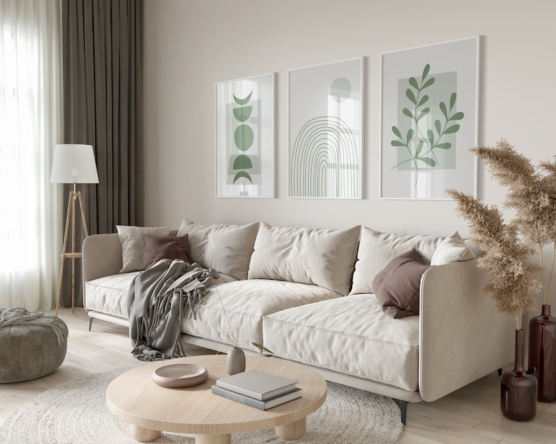 Pu&ograve; includere: Un divano bianco con tre cuscini, una coperta e un tavolino da caff&egrave; rotondo in legno con libri e una ciotola in cima. Il divano si trova in un soggiorno con pareti beige e tre stampe incorniciate con disegni verdi astratti.