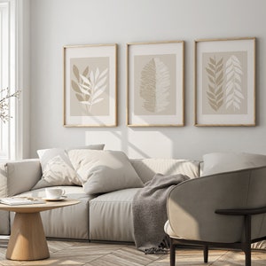 May include: A living room interior with a white sofa, two throw pillows, a throw blanket, a wooden coffee table, and a beige armchair. Three framed prints with abstract leaf designs hang on the wall.