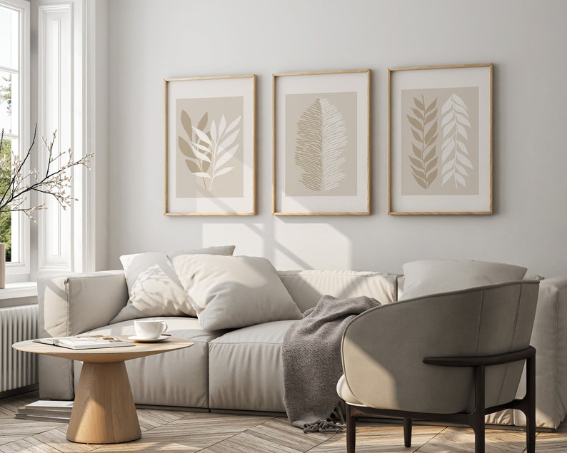 May include: A living room interior with a white sofa, two throw pillows, a throw blanket, a wooden coffee table, and a beige armchair. Three framed prints with abstract leaf designs hang on the wall.