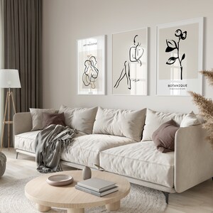 May include: A living room interior with a white sofa, a round coffee table, a lamp, a potted plant, and three framed black and white line art prints on the wall. The prints feature a stylized human figure, a plant, and abstract shapes. The text on the prints reads "Art Moderne" and "Botanique".