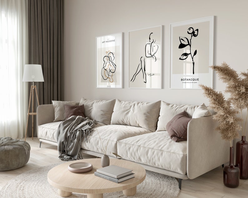 May include: A living room interior with a white sofa, a round coffee table, a lamp, a potted plant, and three framed black and white line art prints on the wall. The prints feature a stylized human figure, a plant, and abstract shapes. The text on the prints reads "Art Moderne" and "Botanique".