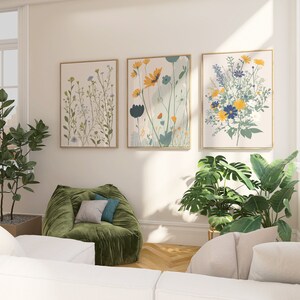 May include: Three framed floral art prints with gold frames hang on a cream-colored wall. The botanical art features colorful flowers in shades of yellow, blue, and green. A green beanbag chair and potted plants are in the foreground.