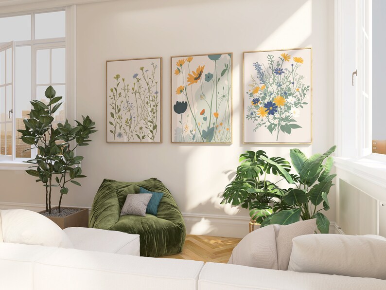 May include: Three framed floral art prints with gold frames hang on a cream-colored wall. The botanical art features colorful flowers in shades of yellow, blue, and green. A green beanbag chair and potted plants are in the foreground.
