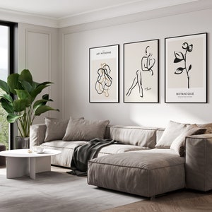 May include: A living room interior with a large sectional sofa in a light beige color. The sofa is positioned in front of a white wall with three framed black and white line art prints. The prints feature abstract shapes, a figure, and a botanical illustration. The text on the prints reads "Art Moderne" and "Botanique". A large potted plant with green leaves is in the corner of the room. A white coffee table with a unique base design sits in front of the sofa. A small gray ottoman is in the foreground.